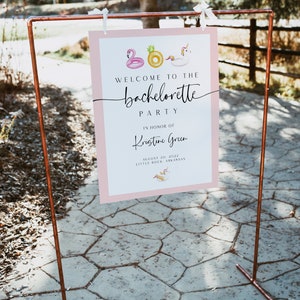 May include: A pink welcome sign with a white border and the text "Welcome to the bachelorette party" in black script font. The sign also includes the text "In honour of Kristine Green" and the date "August 20, 2022". The sign is decorated with small images of inflatable pool toys, including a flamingo, a pineapple, and a unicorn.