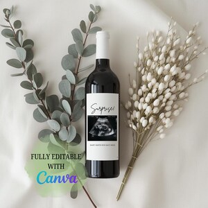 May include: A dark wine bottle with a white label that reads "Surprise!" and features a sonogram image. The bottle is surrounded by eucalyptus and white floral accents, creating a celebratory theme.
