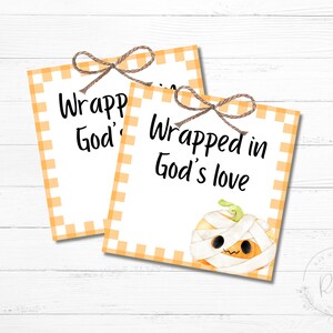 Op de afbeelding: Twee rechthoekige kaarten met oranje ruitjesranden en touwstrikken. De kaarten lezen "Wrapped in God's love" in zwart schrift. Eén kaart bevat een illustratie van een mummiepompoen.