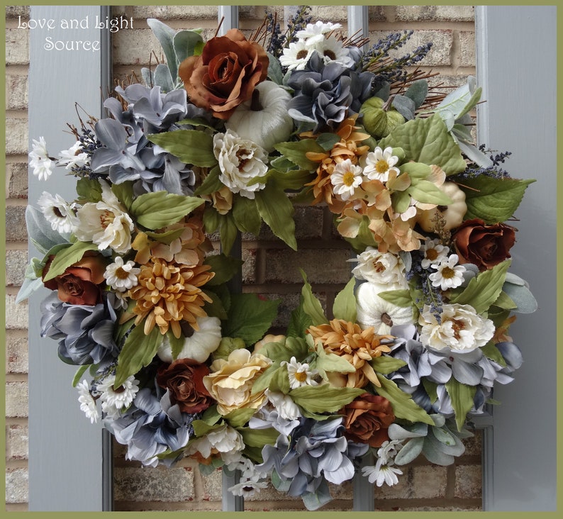 May include: A fall-themed wreath with white pumpkins, brown and white flowers, and blue hydrangeas. The wreath is made of artificial materials and is decorated with green leaves.