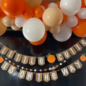 May include: A banner with the text "A little cutie is on the way" with orange and white balloons in the background.