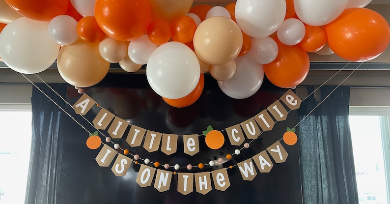 May include: A banner with the text "A little cutie is on the way" with orange and white balloons in the background.