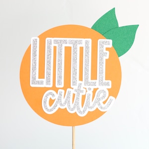 May include: Orange-coloured cake topper with the words "Little cutie" in silver glitter lettering and green leaves on top.