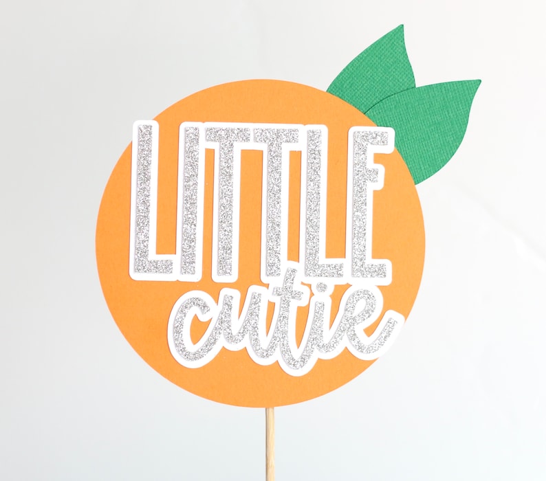 May include: Orange-coloured cake topper with the words "Little cutie" in silver glitter lettering and green leaves on top.