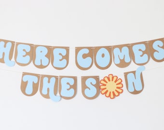 May include: A light blue and brown paper banner with the text "Here Comes The Sun" in a retro font. The sun is depicted with a yellow center and orange rays.