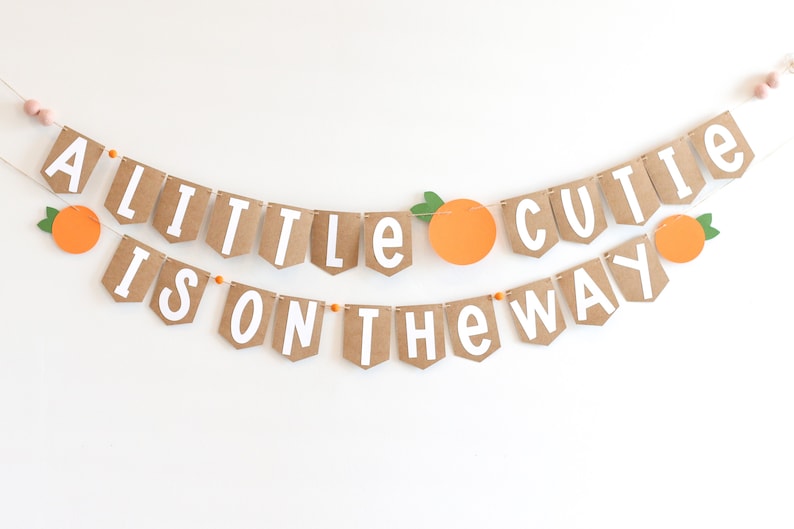May include: A brown paper banner with white letters that reads "A little cutie is on the way". The banner is decorated with orange paper cutouts of oranges.