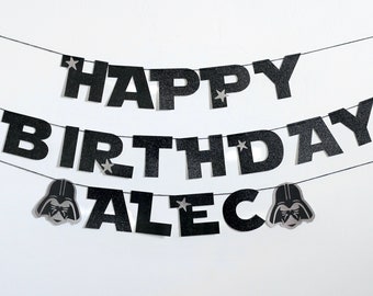 May include: Black glittery banner with the words "HAPPY BIRTHDAY ALEC" in bold, block letters. Silver star accents and Darth Vader head silhouettes add a themed touch. The banner is strung on thin black cords against a white background.