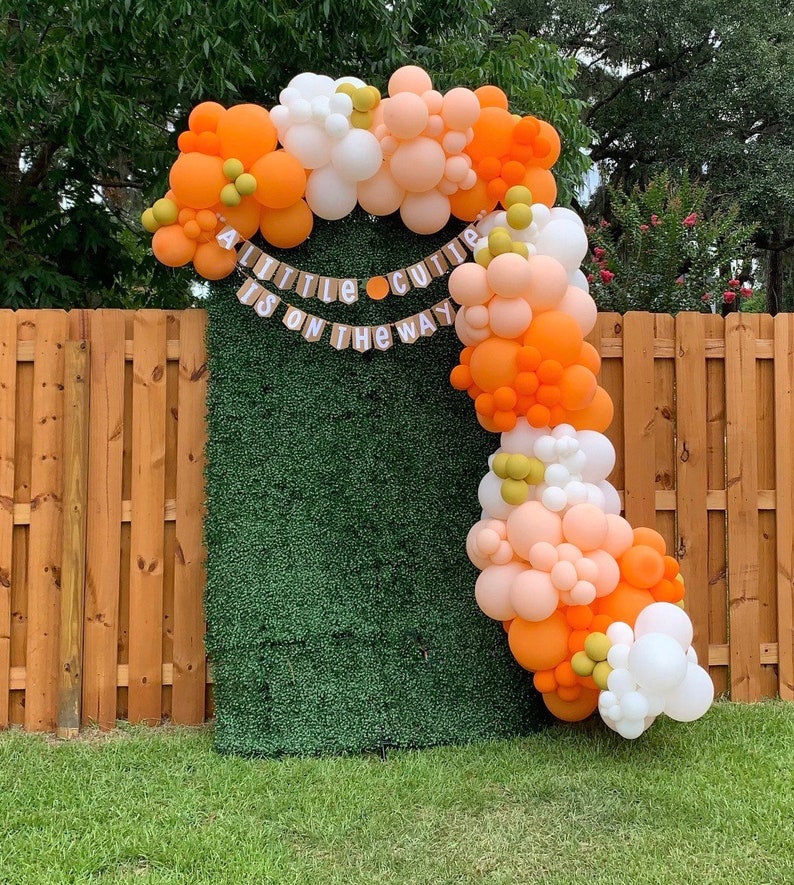 May include: A balloon arch made of orange, peach, white and green balloons. The arch is decorated with a banner that reads "A little cutie is on the way".