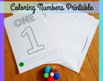 May include: A stack of white coloring pages with the number "1" outlined in black, with the word "ONE" above it. The text "Coloring Numbers Printable" is at the top. A collection of colorful pom-poms sits below the pages.