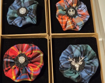 Può includere: Quattro spille in tessuto tartan in scatole individuali. Le spille presentano vari motivi a quadri in rosso, blu e arancione. Ogni spilla ha un centro decorativo, tra cui un gruppo di strass, un emblema argentato e una testa di cervo argentata.