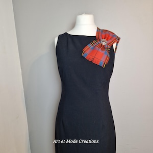 May include: A sleeveless, black dress with a decorative red, blue, and yellow plaid bow pinned to the shoulder. The bow features a silver-colored brooch in the center. The text "Art et Mode Creations" is visible at the bottom of the image.