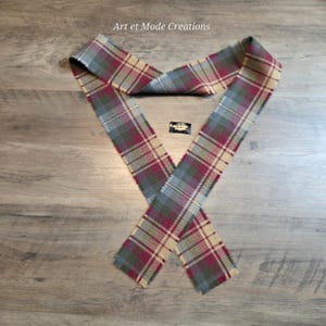 Puede incluir: Una bufanda a cuadros en tonos burdeos, grises y beige, dispuesta en forma de V. La bufanda presenta un diseño de tartán clásico. El texto "Art et Mode Creations" es visible en la parte superior. También hay una pequeña etiqueta.