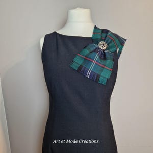 May include: A black sleeveless dress with a green, blue, and red tartan bow pinned to the shoulder. The bow is secured with a silver brooch. The text "Art et Mode Creations" is at the bottom.