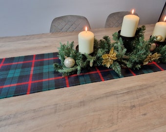 May include: A wooden table with a plaid table runner and a centerpiece of evergreen branches, gold ornaments, and three white pillar candles.