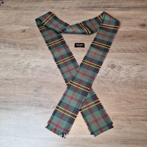 May include: A green, red, yellow, and navy blue plaid scarf with frayed edges. The scarf is in an X shape on a wooden surface. The text "Art et Mode Creations" is visible on a small black label.