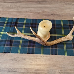 Puede incluir: Un portavelas de cuerno de ciervo natural con una vela de color crema sobre una base de madera. El cuerno descansa sobre un camino de mesa a cuadros azul, verde y negro. El fondo es una superficie de madera clara.