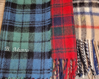 May include: Three plaid scarves with fringed edges. The scarf on the left is blue, green, and black with the text "B. Munro" embroidered on it. The middle scarf is red, black, and blue. The scarf on the right is beige, black, and white.