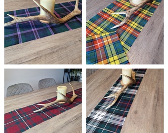 May include: Four table runners with different tartan patterns. Each runner is displayed on a wooden table with a decorative antler and a candle. The table runners are in shades of green, red, yellow, and blue.