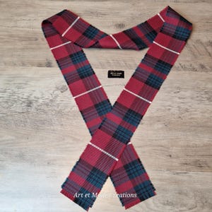 May include: A red, black, white, and teal tartan scarf. The scarf is made of woven fabric and features a classic plaid pattern. The scarf is displayed on a wooden surface. The text "Art et Mode Creations" is visible.