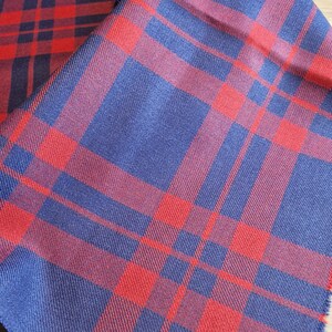 May include: Red and blue plaid fabric with a fringed edge. The fabric has a woven texture and is folded in half.