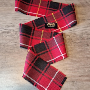 May include: Red, black and white plaid ribbon with frayed edges. The ribbon is folded and arranged on a wooden surface. The label "Art et Mode Creations" is visible on the ribbon.