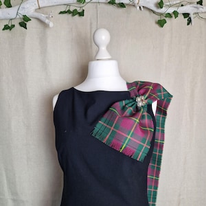 May include: A black sleeveless dress with a tartan sash draped over the shoulder. The sash features a red, green, and yellow plaid pattern, secured with a decorative brooch. The dress is displayed on a white mannequin.