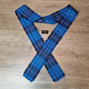 May include: A blue tartan scarf with black and pink lines, displayed on a wooden surface. The scarf has a fringed edge and the text "Art et Mode Creations" is visible.