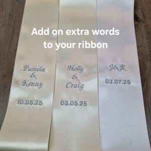 May include: Cream satin ribbons with embroidered names and dates. The ribbons show text such as "Pamela & Kenny 10.05.25", "Holly & Craig 03.05.25", and "J&R 03.07.25". The phrase "Add on extra words to your ribbon" is also displayed.