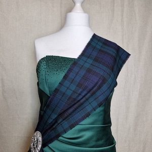 Peut inclure: Une robe en satin vert avec un sash en plaid drapé sur une épaule. Le sash est sécurisé par une broche argentée. La robe est sans bretelles et a un corsage ajusté.