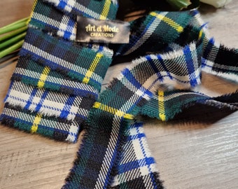 May include: A close-up of a tartan ribbon with frayed edges. The ribbon features a pattern of green, blue, yellow, and white stripes. A small tag with the text "Art et Mode CREATIONS" is visible. The ribbon is lying on a wooden surface.