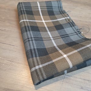 May include: Folded fabric with a plaid pattern in shades of gray, brown, and white. The fabric has a woven texture and a frayed edge. The plaid design features intersecting lines creating a classic tartan look.