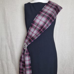 Peut inclure: Une robe noire sans manches avec une écharpe à carreaux drapé sur une épaule. L'écharpe est nouée à la taille et comporte une épingle décorative.