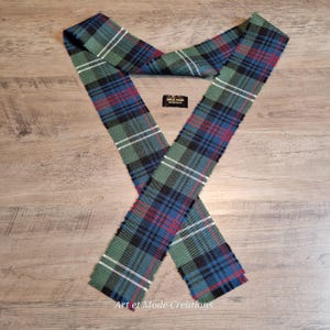 May include: A tartan scarf in green, blue, black, and red, arranged in an X shape. The text "Art et Mode Creations" is visible on a small black tag and at the bottom of the scarf.