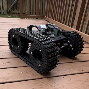 Peut inclure: Robot noir télécommandé avec des chenilles de char et une caméra. Le robot a une poignée sur le dessus et est posé sur une terrasse en bois. Les chenilles du robot sont faites de pièces imbriquées.