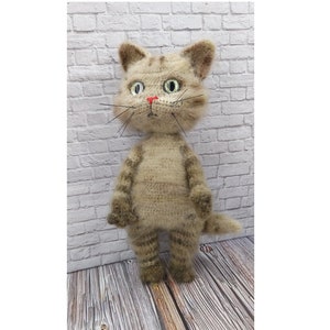 May include: A handmade cat doll in light brown and tan hues, standing upright. It features green eyes, a red nose, and black whiskers. The doll is crafted from a soft, textured material and is set against a white brick wall background.