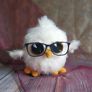 Puede incluir: Un búho blanco de crochet que lleva gafas de montura azul con ojos negros y blancos. El búho tiene patas de crochet amarillas y está sentado sobre un fondo de tela burdeos.