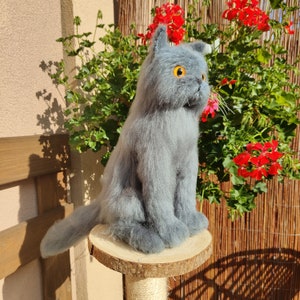 Può includere: Un giocattolo di gatto di peluche grigio seduto su un trespolo per gatti in legno. Il gatto ha gli occhi arancioni e una coda soffice.