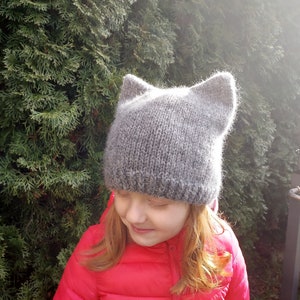 May include: A young person wearing a gray knitted hat with cat ears and a red puffer jacket.