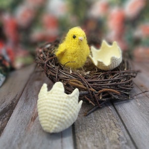 Pode incluir: Um pintinho amarelo de malha senta-se num ninho de ramos marrons com duas cascas de ovo de crochê branco. Uma casca de ovo está partida.