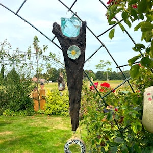 Könnte beinhalten: Ein rustikaler Holzast mit einem blauen Glasstück an der Spitze, einer weißen und gelben Keramikblume und einem Keramikring mit einem schwarz-weißen Muster, der daran hängt.