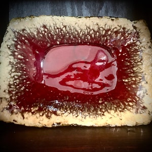 Może przedstawiać: Prostokątne ceramiczne naczynie z czerwoną glazurą i fakturowanym, beżowym brzegiem. Glazura ma błyszczące wykończenie i tworzy unikalny, abstrakcyjny wz&oacute;r.