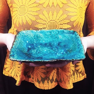 Może przedstawiać: Kwadratowa ceramiczna miska z turkusową glazurą i szorstkim, fakturowanym brzegiem. Miska jest trzymana przez dłonie osoby.