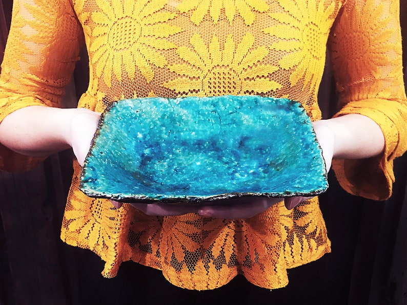 Może przedstawiać: Kwadratowa ceramiczna miska z turkusową glazurą i szorstkim, fakturowanym brzegiem. Miska jest trzymana przez dłonie osoby.