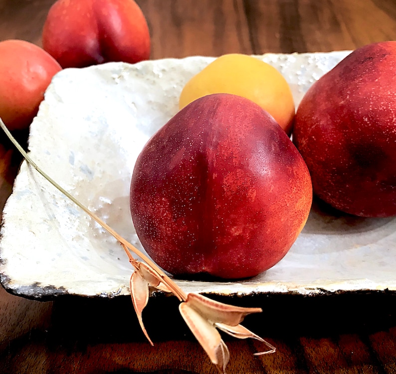 Może przedstawiać: Biała ceramiczna miska z czterema brzoskwiniami. Dwie brzoskwinie są czerwono-ż&oacute;łte, a dwie są czerwone. Brzoskwinie są ułożone w p&oacute;łkole.