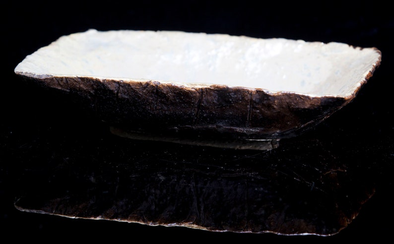 Może przedstawiać: Prostokątne ceramiczne naczynie z białym wnętrzem i ciemnobrązowym zewnętrzem. Naczynie ma szorstką, fakturowaną powierzchnię i wydaje się być wykonane ręcznie.