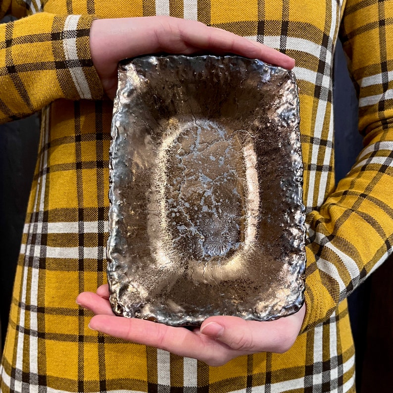 Może przedstawiać: Prostokątne ceramiczne naczynie z fakturowanym, metalicznym wykończeniem. Naczynie ma lekko uniesiony brzeg i gładkie, owalne wnętrze.