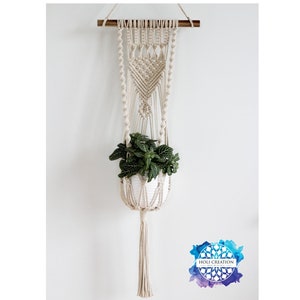 Puede incluir: Un colgador de plantas de macramé con una maceta blanca que contiene una planta verde. El colgador de color beige está suspendido de una varilla de madera. El diseño presenta intrincados patrones de nudos y una larga borla en la parte inferior. El fondo es una pared blanca lisa.