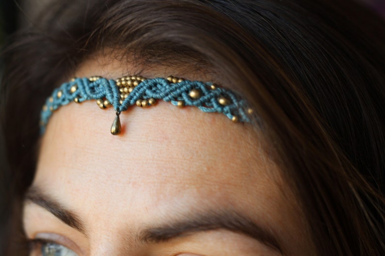 May include: A teal blue and gold beaded headband with a teardrop pendant. The headband is worn on a person's forehead.