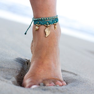 May include: A teal and gold ankle bracelet with leaf-shaped charms. The bracelet is worn on a person's ankle on a sandy beach.
