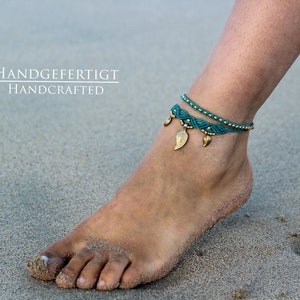 May include: A teal blue and gold ankle bracelet with leaf charms. The bracelet is handmade and the text "HANDGEFERTIGT HANDCRAFTED" is visible in the image.